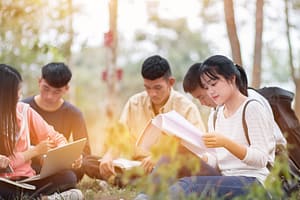 giáo dục học tập nghiên cứu khái niệm ngoài trời: nhóm sinh viên châu Á tự tin ngồi đọc sách, sử dụng máy tính bảng trên bãi cỏ xanh trong trường đại học với nụ cười hạnh phúc.